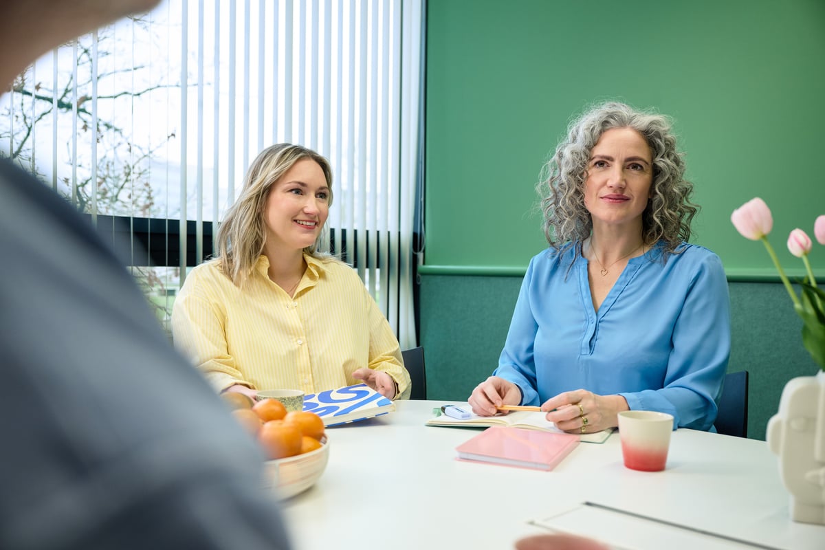 Two women are sitting at a table in a brightly lit room with a green wall, smiling and talking.