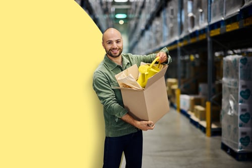 Een glimlachende man in een magazijn, gekleed in een groen shirt, houdt een kartonnen doos met een gele gieter vast.