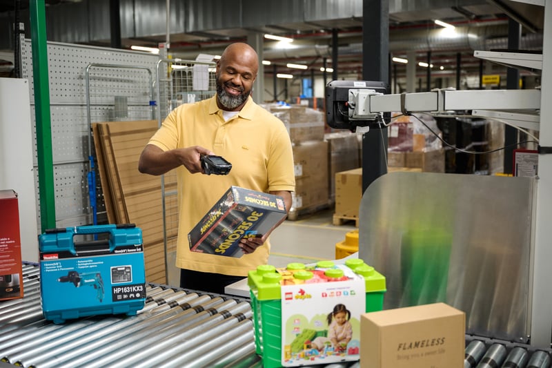 A man in a yellow shirt smilingly scans a box on a conveyor belt in a warehouse. Nearby are toolboxes and toy packaging.
