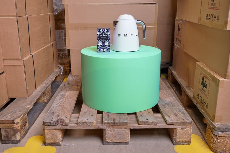 Op een ronde groene pilaar die op een houten pallet staat, staan een witte waterkoker en een doos thee, omringd door kartonnen dozen in een magazijnomgeving.