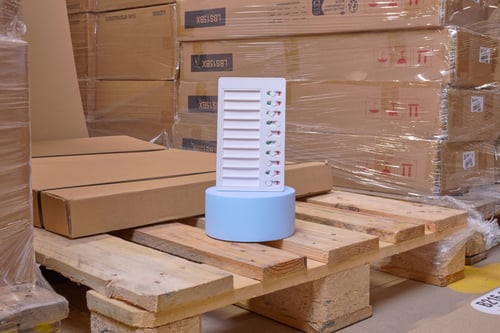 Een blauwe standaard met een checklist erop staat op een houten pallet, omringd door kartonnen dozen.