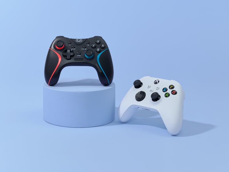 Twee gamecontrollers staan tegen een lichtblauwe achtergrond: een zwarte controller met rode en blauwe accenten op een voetstuk en daarnaast een witte controller.