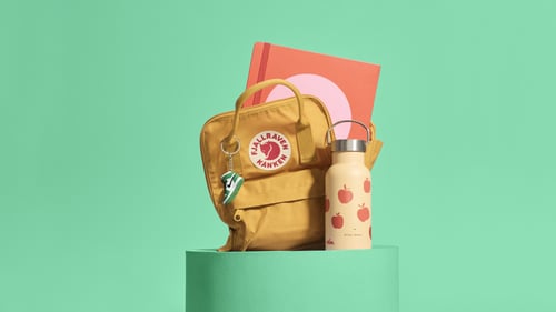 A mustard yellow backpack and an apple-patterned water bottle stand on a green pedestal. Behind them, a pink and red notebook is visible. The background is green.