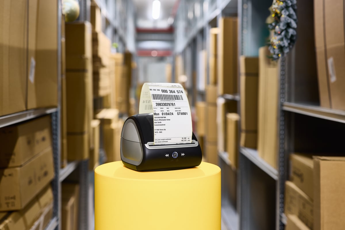 Een labelprinter staat op een geel voetstuk in een magazijn, omringd door schappen vol kartonnen dozen. De omgeving is georganiseerd en efficiënt.