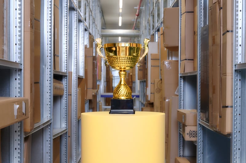 A golden trophy on a yellow pedestal is centered in an aisle with tall shelves full of cardboard boxes, radiating a sense of achievement and focus.
