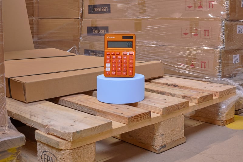 Une scène d'entrepôt avec des cartons empilés sur des palettes en bois. Dessus se trouve un cylindre bleu avec une calculatrice orange posée dessus.