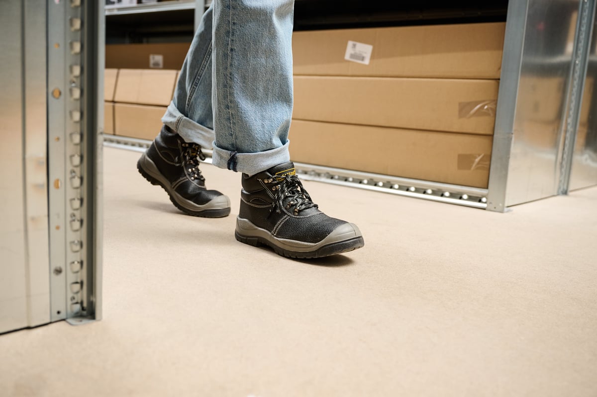 Close-up van een persoon die zwarte werklaarzen en opgerolde spijkerbroek draagt en langs metalen rekken in een magazijn loopt.