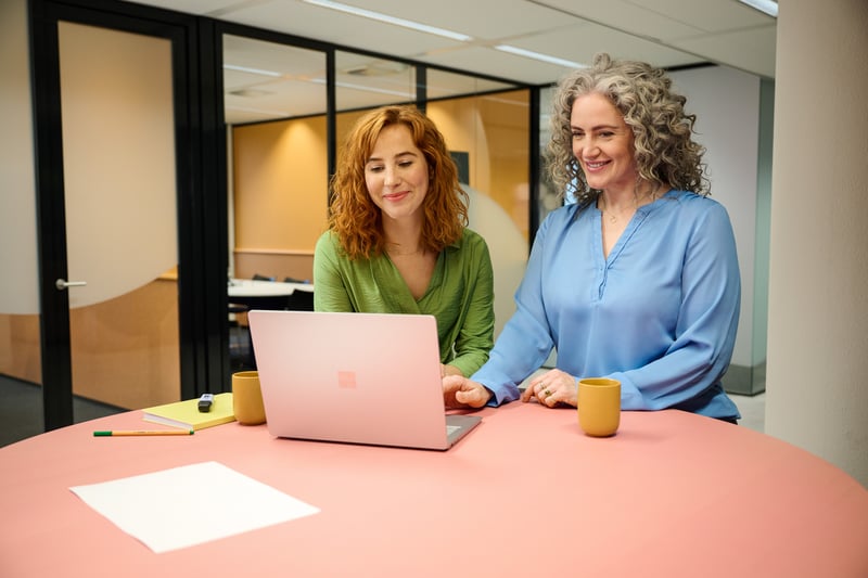 Two women stand together behind a laptop.