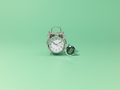 Twee metalen klokken, een grote en een kleine, staan op een mintgroene achtergrond. De grote klok geeft 10:10 aan, terwijl de kleine een digitaal display heeft.