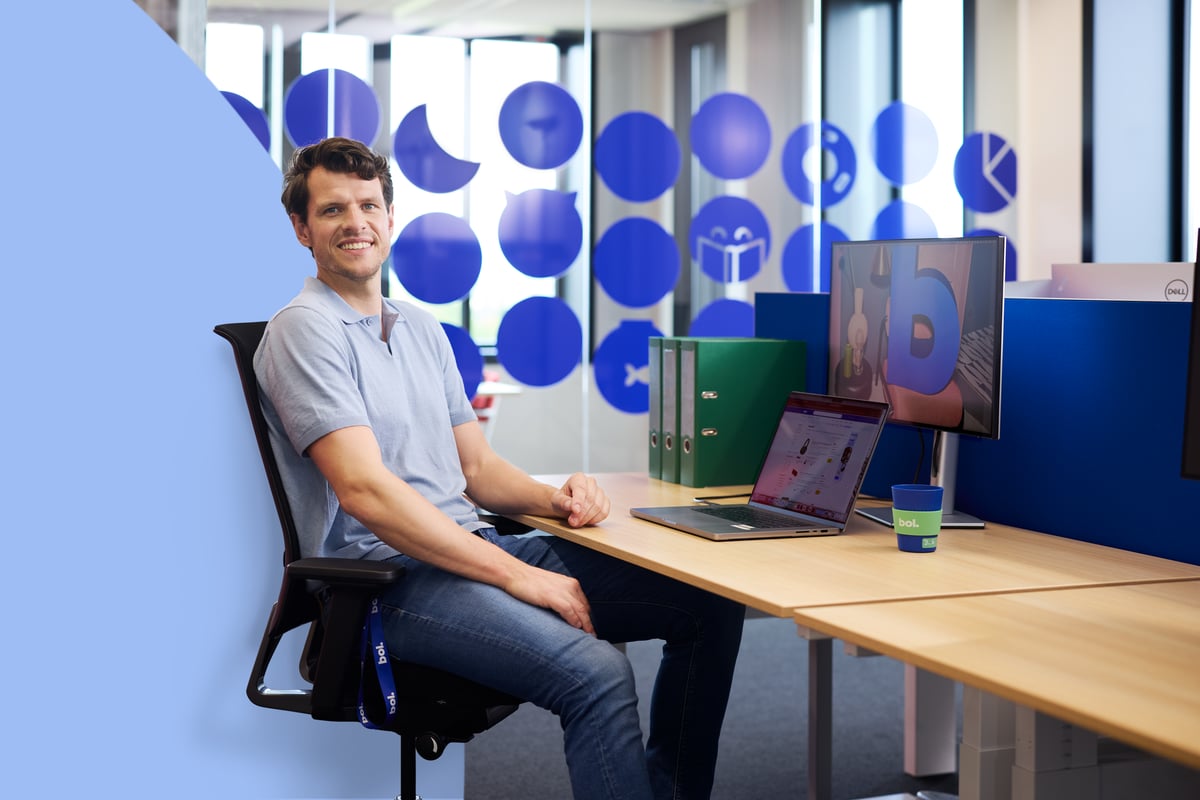 Ein Mann in einem hellblauen Hemd sitzt lächelnd an einem Schreibtisch mit einem Laptop und einem Monitor. Die Büroumgebung ist hell, mit blauer Beschriftung an der Glaswand.