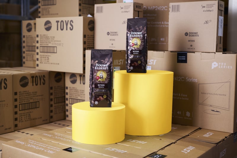Twee koffiezakken staan op gele cilindervormige standaards tussen gestapelde kartonnen dozen op een houten pallet, wat een industriële en levendige presentatie creëert.