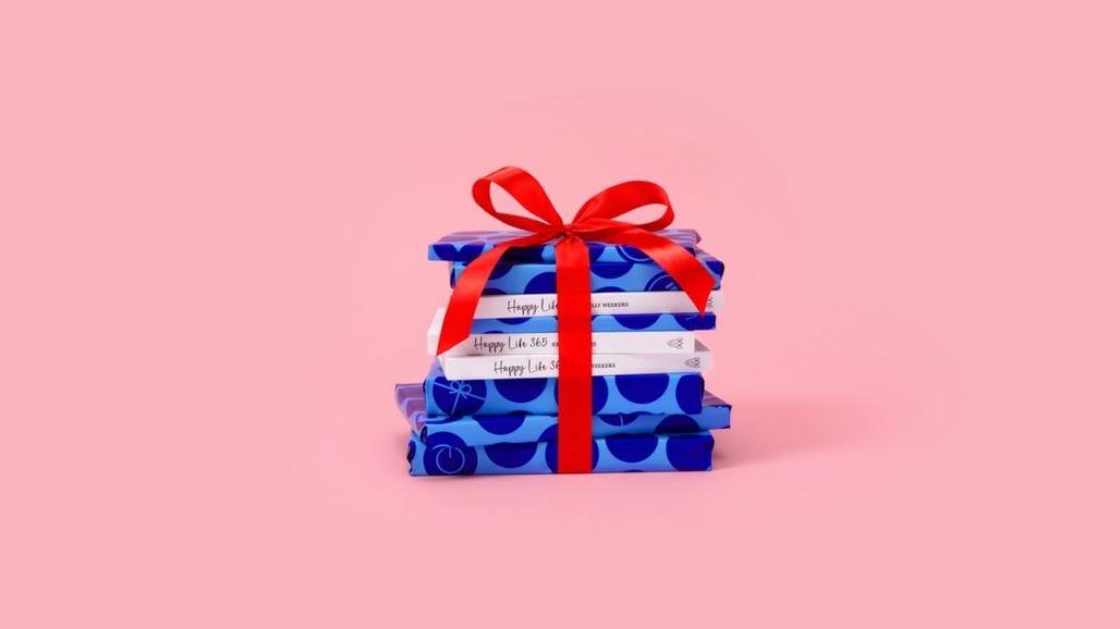 A stack of notebooks, wrapped in vibrant blue with various patterns, is tied with a bright red ribbon against a soft pink background