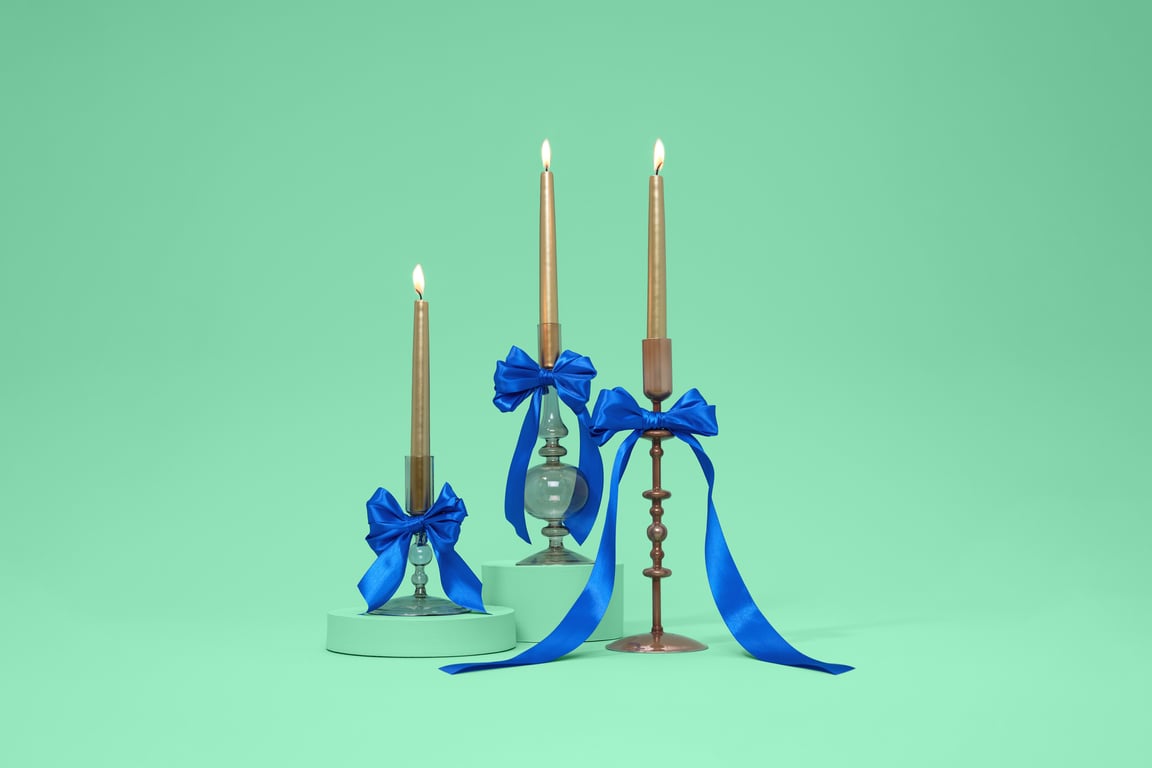 Three lit candles in elegant holders adorned with vibrant blue bows against a soft green background