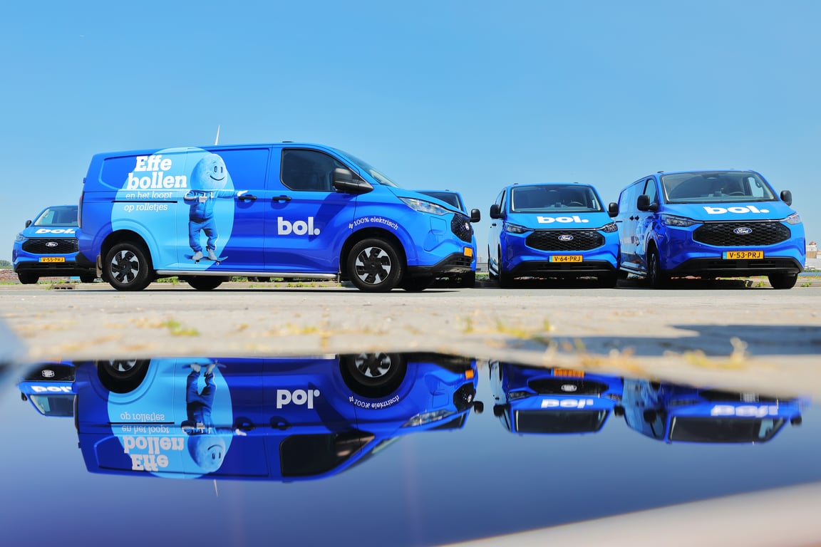 A blue Ampère van parked by the side of the road, surrounded by greenery and residential buildings.
