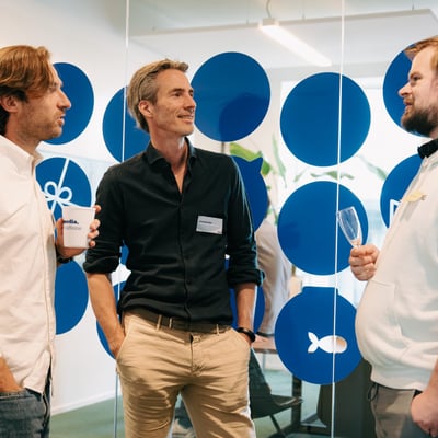 Drie mannen praten met elkaar in een modern kantoor, staand bij een glazen wand versierd met grote blauwe cirkels.