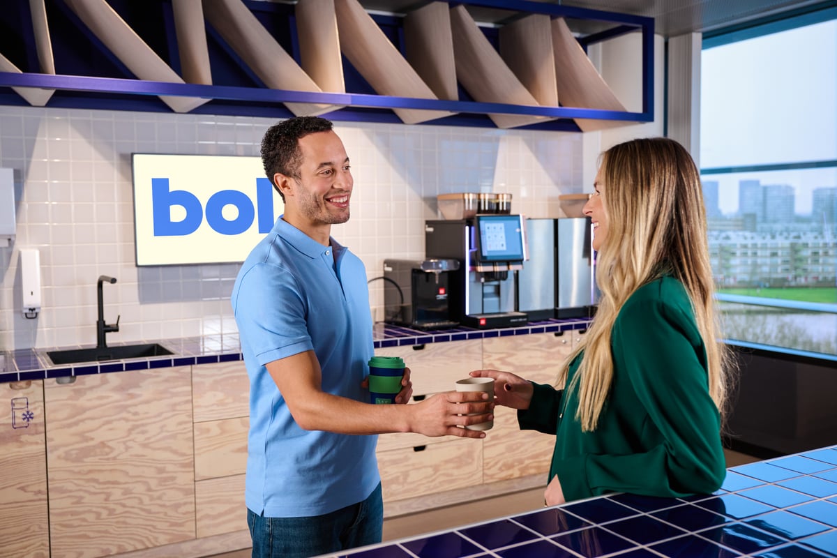 Een man en een vrouw glimlachen, de man geeft de vrouw een kop koffie in een moderne kantoorkeuken, met een blauw-witte betegelde aanrecht, een koffiezetapparaat en uitzicht naar buiten.