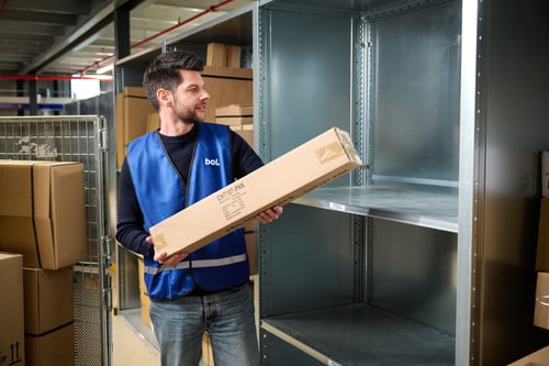 A warehouse worker in a blue vest holds a rectangular package, looking at empty metal shelves, with cardboard boxes in the background. The mood is busy and focused.