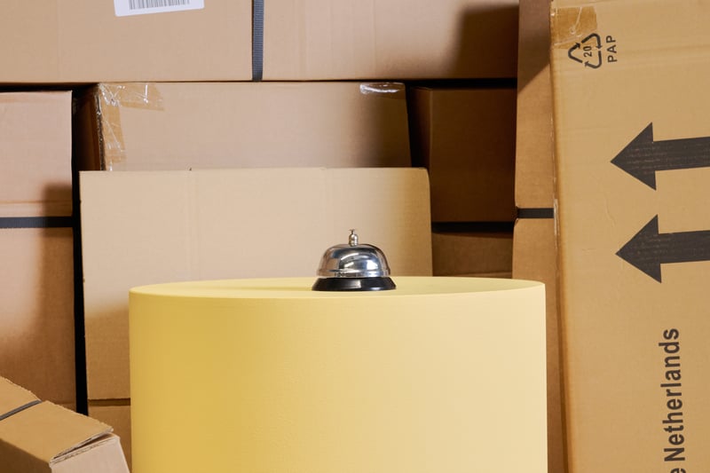 A silver service bell stands on a large yellow cylindrical object, surrounded by stacked cardboard boxes.