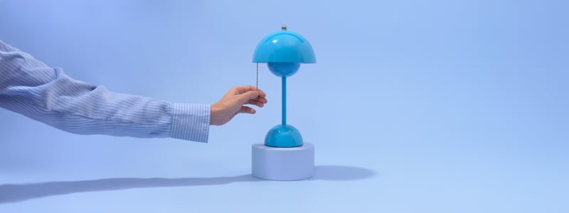Een hand trekt aan het kettinkje van een kleine, moderne blauwe lamp op een witte voet, tegen een blauwe achtergrond.