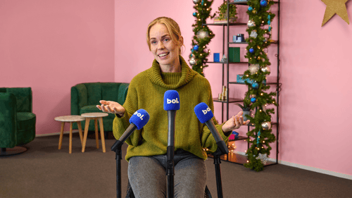 Een vrouw in een groene trui spreekt vol vertrouwen in meerdere blauwe microfoons met ‘bol.’. Ze staat in een feestelijke ruimte met roze muren en kerstdecoratie.