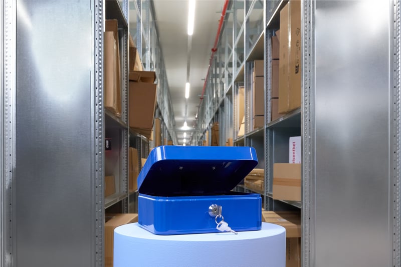 A blue safe with keys stands open on a pedestal in a warehouse aisle, surrounded by shelves with boxes.