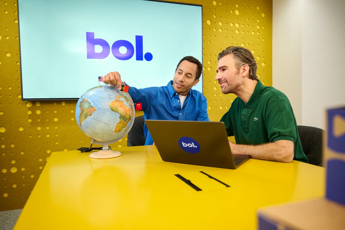 Two men at a yellow table, one points to a globe, the other uses a laptop.